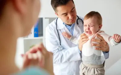 Lekarz trzyma płaczące dziecko, a z przodu widać zamazaną postać matki.