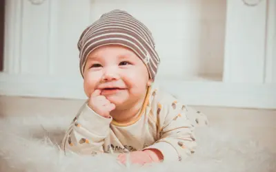 Uśmiechnięte dziecko w czapce leży na białym futerku, z palcem w ustach.