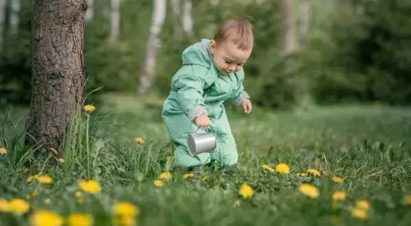 Małe dziecko w zielonym kombinezonie bawi się konewką wśród traw i żółtych kwiatów.